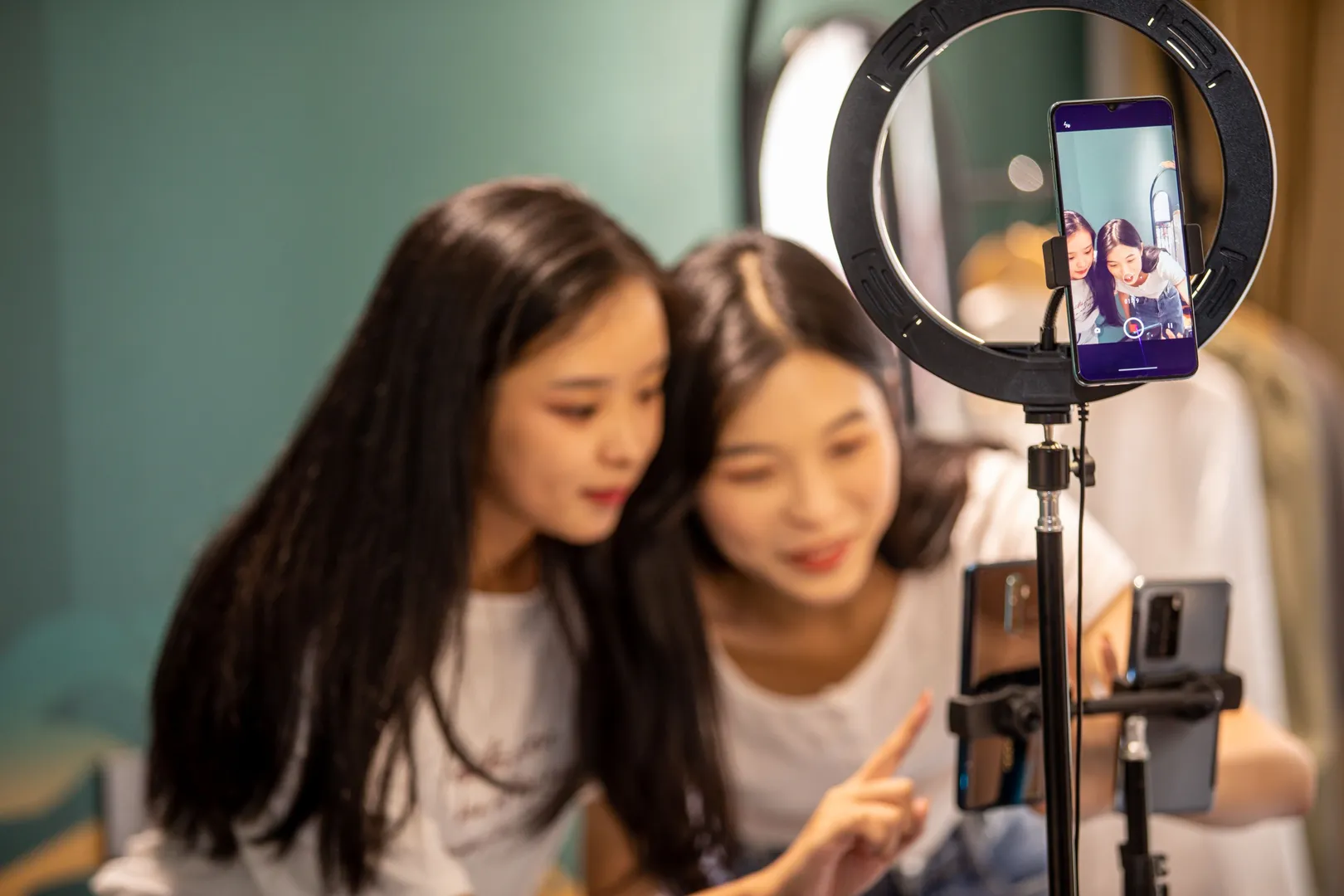 Two women huddled around a mobile phone reviewing filmed content beside a ring light.