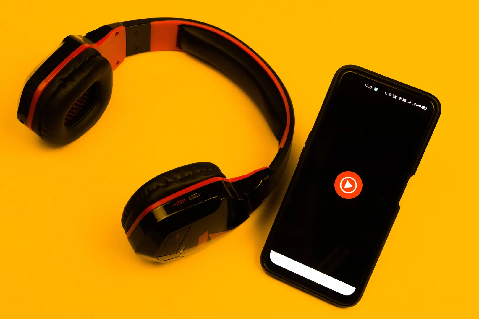 Black and red headphones beside a smartphone displaying a video app on a bright yellow background.