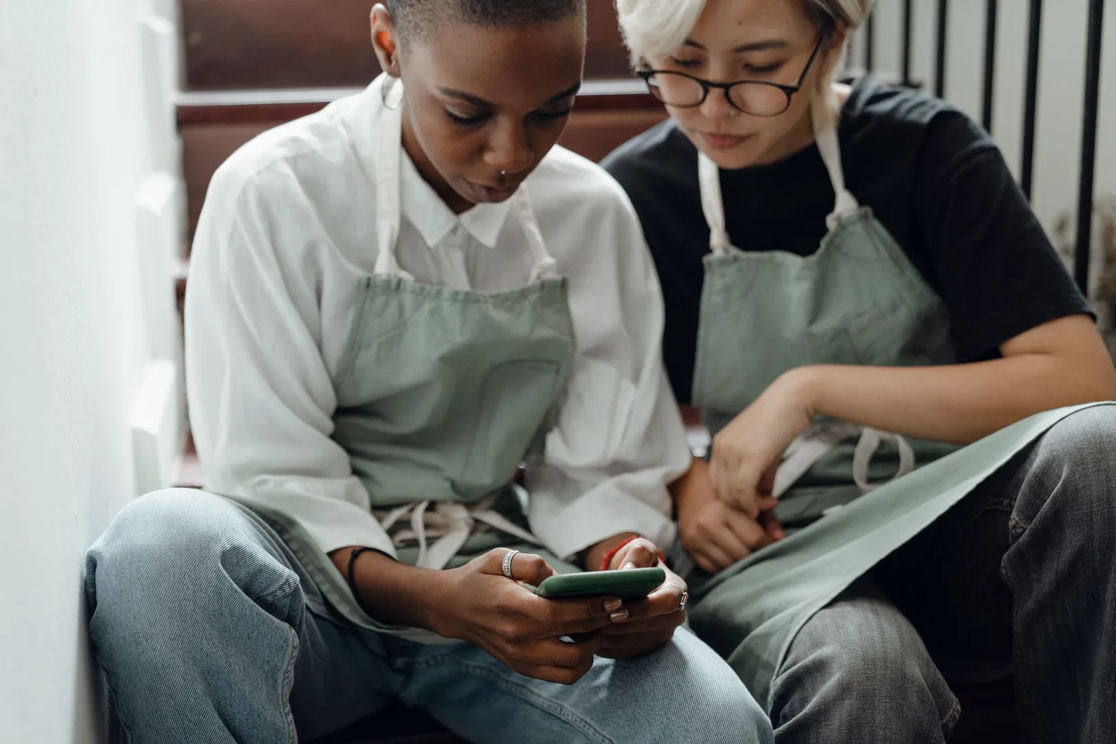 wo people wearing cooking aprons sit on stairs while looking at a smartphone together.