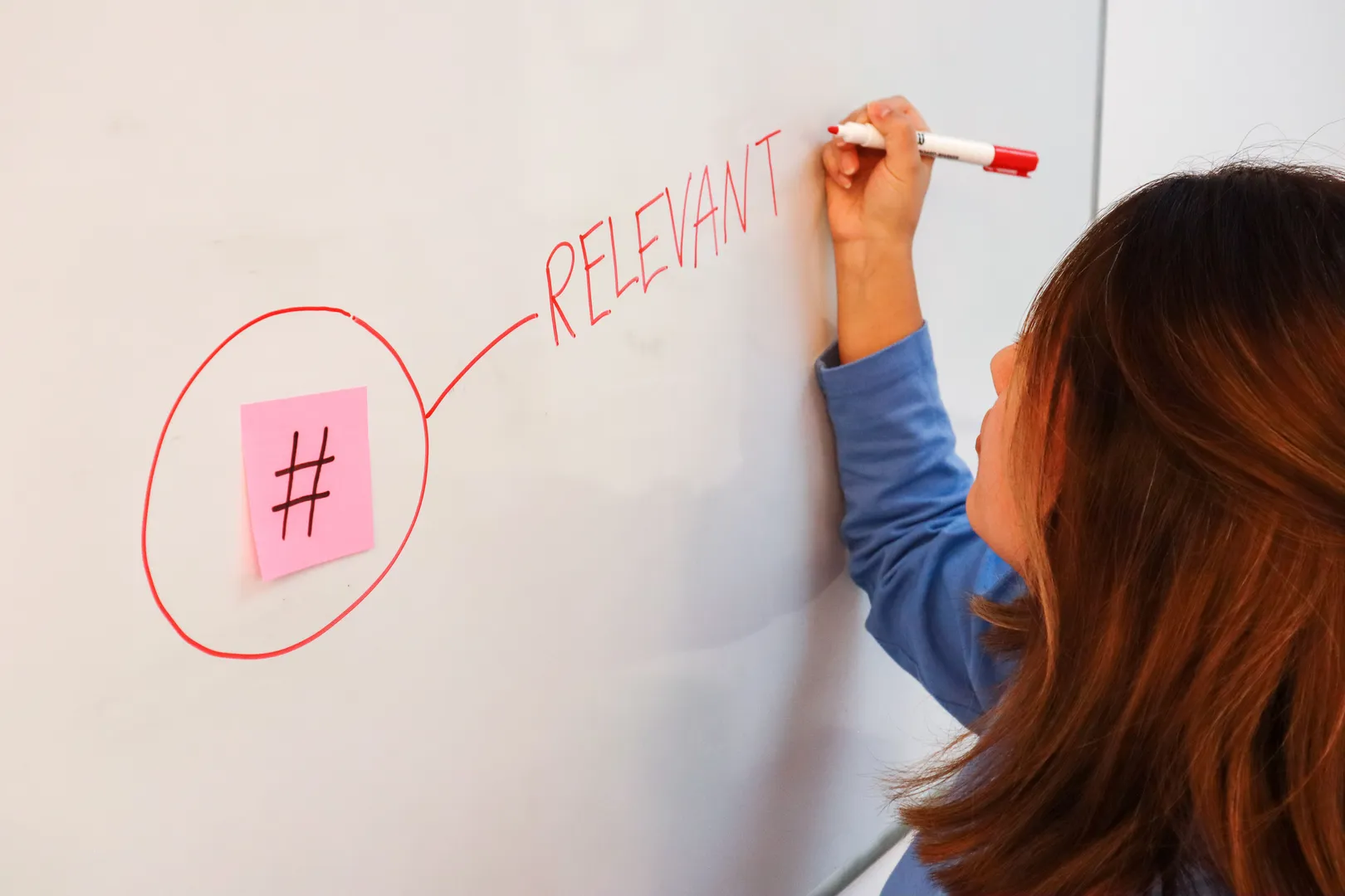 Woman writing “relevant” in red on a whiteboard beside a circled sticky note featuring a hashtag symbol.